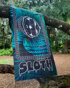 SLOTH Jungle Pashmina draped over a tree branch: turquoise palm print with the SLOTH character, chain border and drippy SLOTH lettering