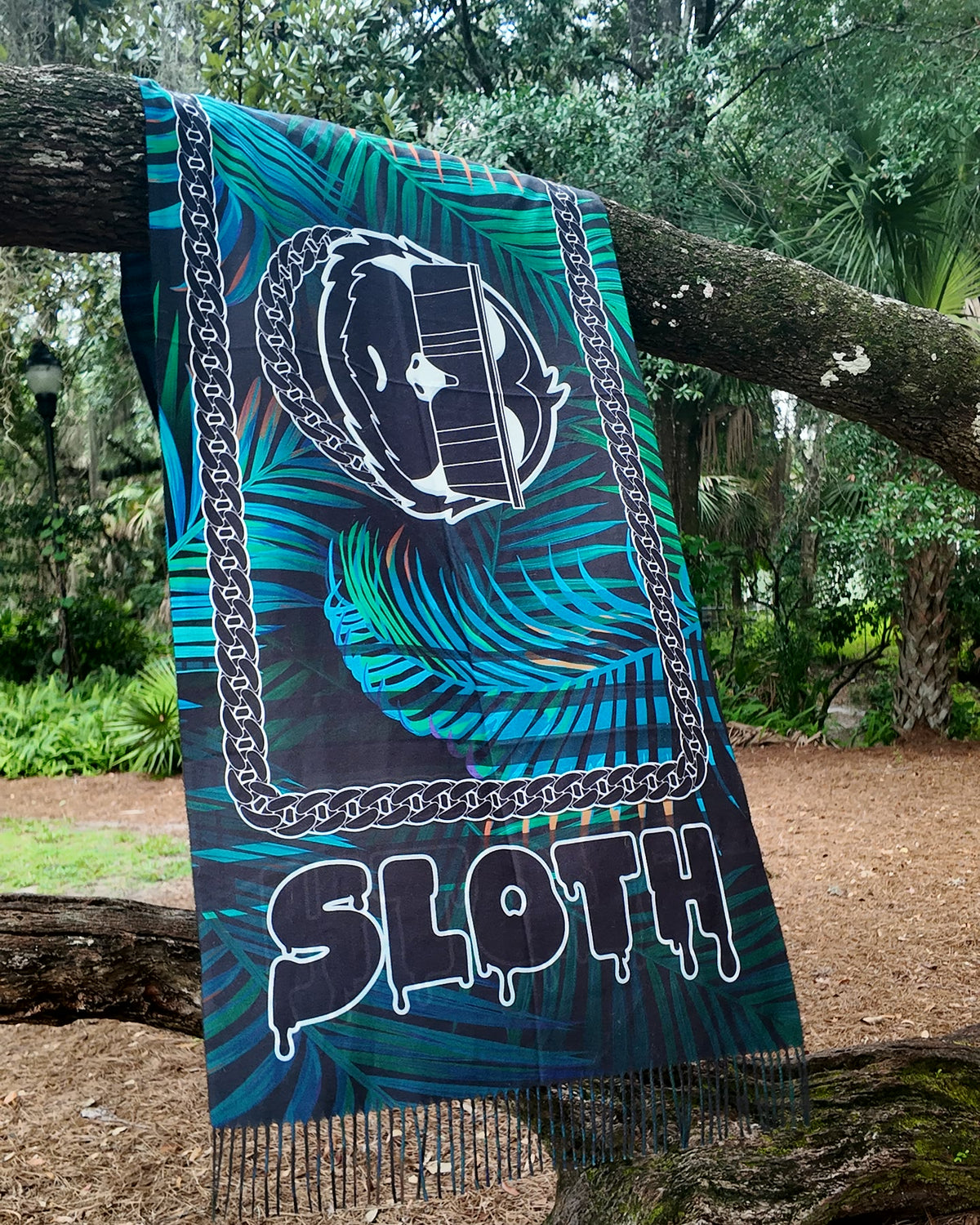 SLOTH Jungle Pashmina draped over a tree branch: turquoise palm print with the SLOTH character, chain border and drippy SLOTH lettering